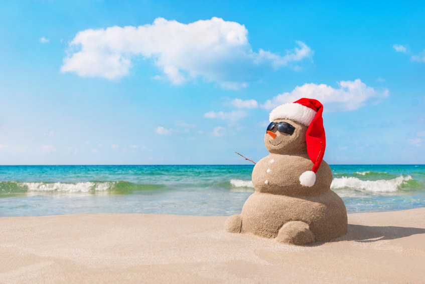 Sandy christmas snowman in red santa hat and sunglasses at sunny beach. Holiday concept for New Years Cards.
