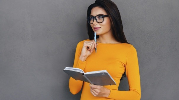 Waiting for inspiration. Thoughtful young and beautiful woman touching chin with pen and holding notebook while standing against grey background