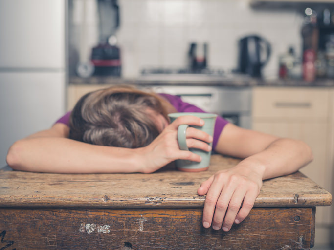 A tired young woman is having a cup of tea and is resting her head on a table
